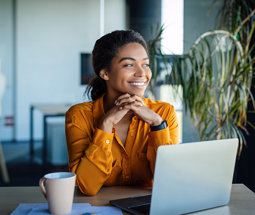 A woman in a yellow blouse sits at her laptop researching 2024 cybersecurity predicitons with a cup of coffee looking out the window.
